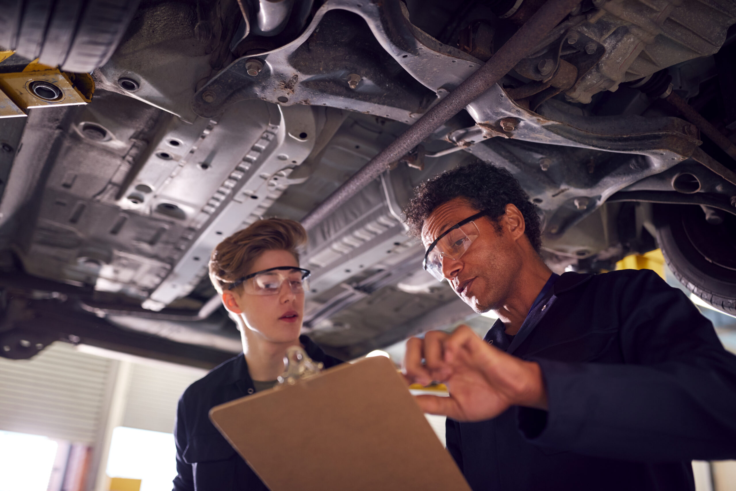 Male Tutor With Student Looking Underneath Car On Hydraulic Ramp On Auto Mechanic Course